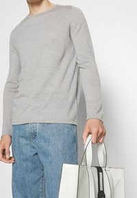 Grå långärmad tröja i mjukt material, kombinerad med ljusblå jeans. En vit axelväska med svarta detaljer hålls.