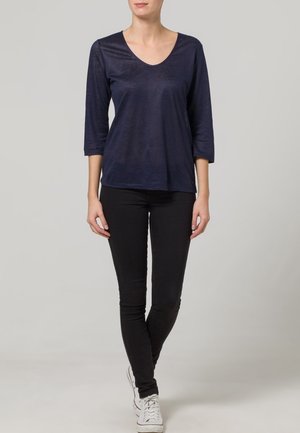 Žena stojící se zkříženýma nohama, oblečená v tmavě modrém dlouhém tričku, černých kalhotách a bílých teniskách na světle šedém pozadí.