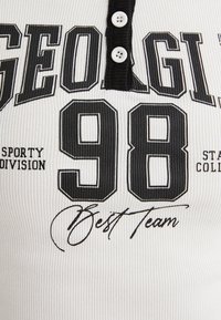 Polo shirt bianco a coste con bottoni neri, numeri neri sovradimensionati "98" e testo "GEORGIA SPORTY DIVISION" con accento in corsivo.