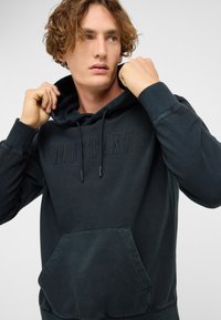 Tmavozelená mikina s kapucňou z mäkkej tkaniny, vybavená klokáním vreckom, nastaviteľnými šnúrkami a reliéfnym logom "MUSTANG" na hrudi.