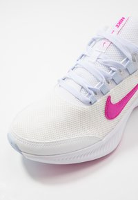 Bílé síťované sportovní boty s světle modrými akcenty a výrazným růžovým logem swoosh. Vybaveny texturovaným povrchem a šněrovacím designem.