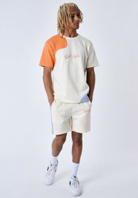 Camiseta de colores claros, de manga corta, con acentos en naranja y azul; combinada con pantalones cortos a juego. Ambos presentan texto bordado. Se llevan zapatillas deportivas.