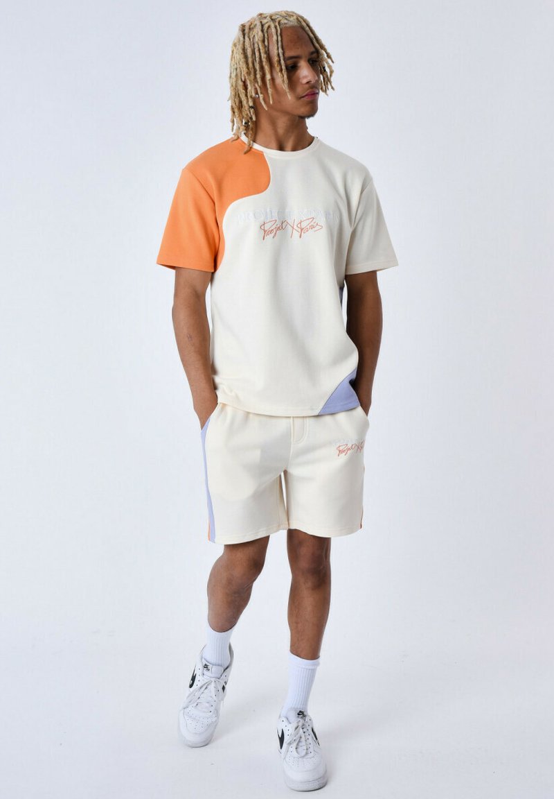 Camiseta de colores claros, de manga corta, con acentos en naranja y azul; combinada con pantalones cortos a juego. Ambos presentan texto bordado. Se llevan zapatillas deportivas.