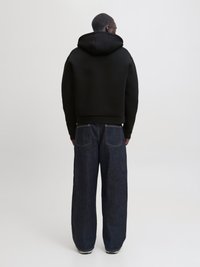 Άνδρας με την πλάτη γυρισμένη φοράει μαύρο φούτερ με κουκούλα και φαρδιά σκούρα μπλε τζιν με λευκή ραφή, στέκεται μπροστά σε μια απλή ανοιχτογκρι φόντο.