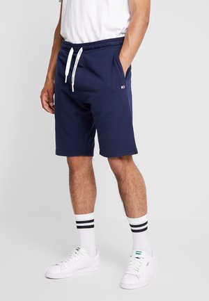 Shorts en coton bleu marine avec cordon blanc, poches latérales et petit logo ; associés à des baskets blanches et des chaussettes rayées.