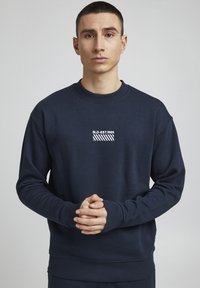 Sudadera navy en mezcla de algodón, cuello redondo, puños y dobladillo de canalé, presenta texto gráfico blanco y un patrón a rayas en el pecho.