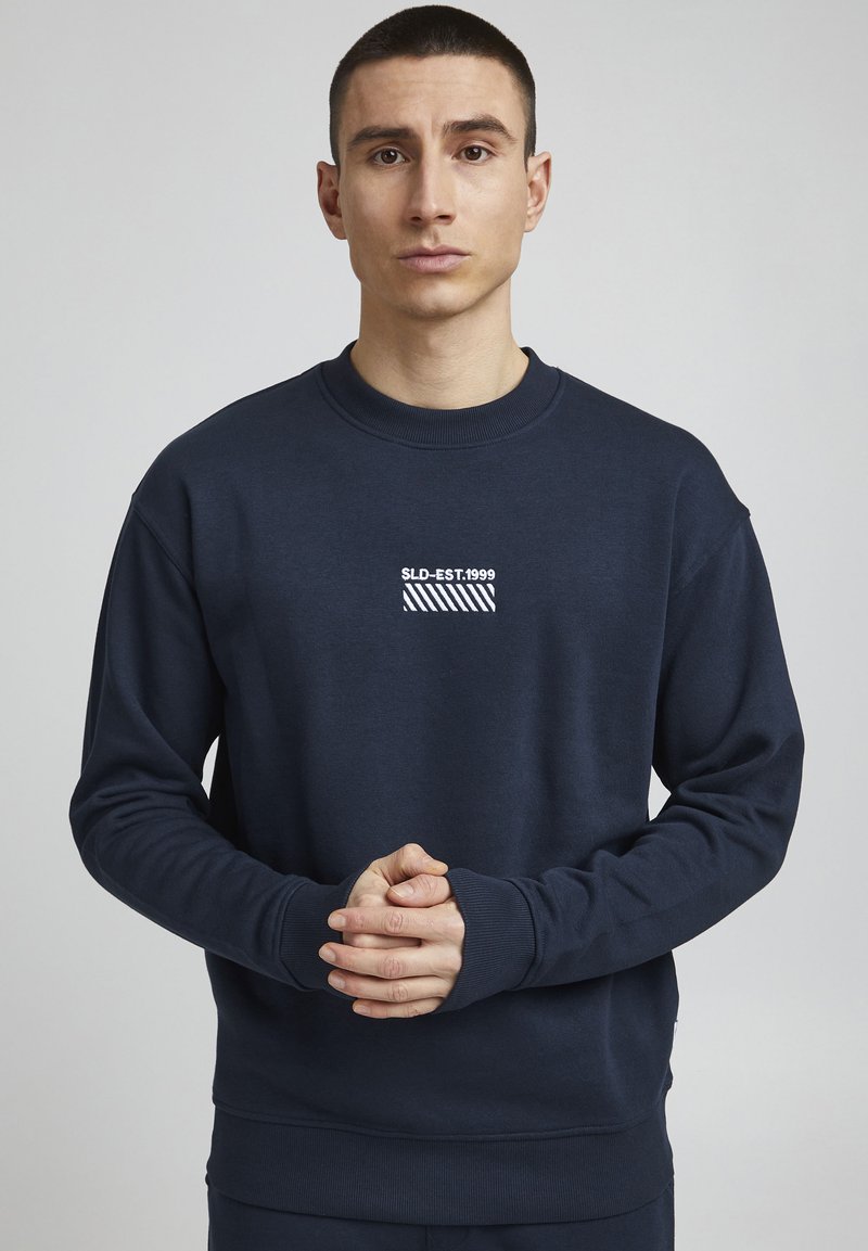 Sudadera navy en mezcla de algodón, cuello redondo, puños y dobladillo de canalé, presenta texto gráfico blanco y un patrón a rayas en el pecho.