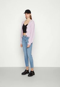 Cardigã lilás de malha com grandes botões pretos, usado sobre um top preto curto, combinado com jeans skinny azul claro e sandálias pretas.