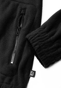Manche et poche d'une veste polaire noire avec fermeture éclair et une petite étiquette comportant un logo stylisé « B » sur la couture latérale.