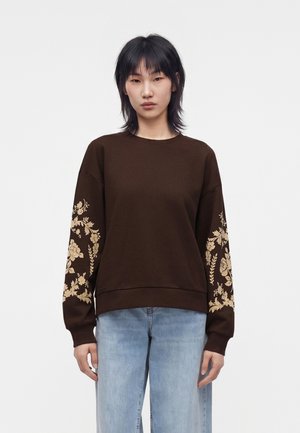 Jeune femme portant un sweat-shirt marron avec des motifs floraux beiges sur les manches et un jean bleu clair, debout devant un fond uni.