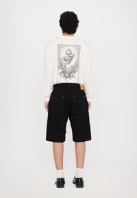 Cămașă albă cu mânecă lungă, având un grafic negru pe spate, asortată cu pantaloni scurți negri și încălțăminte neagră, creând un aspect contrastant.