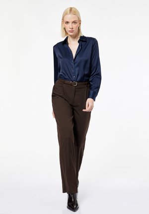 Mujer rubia con camisa de satén azul marino abotonada, pantalones marrones de cintura alta con cinturón y botas negras puntiagudas, caminando hacia la cámara.