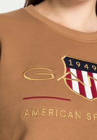 Brauner Sweatshirt mit geripptem Halsauschnitt, gesticktem Logo mit Schild-Design und "GANT" in goldenen und roten Akzenten.