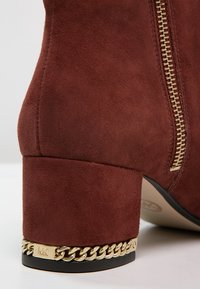 Gros plan sur un talon de botte en suède marron avec un détail de chaîne dorée et une fermeture éclair sur le côté.