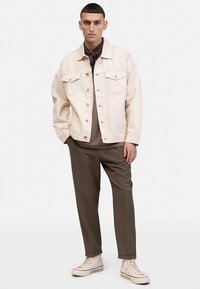 Lichtbeige denimjack met borstzakken, knopen en een ontspannen pasvorm, gecombineerd met donkerbruine broek en witte sneakers.