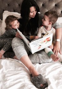 Una madre e due bambini siedono su un letto, leggendo un vivace libro per bambini. Indossano pigiami grigi e pantofole di peluche su lenzuola morbide.