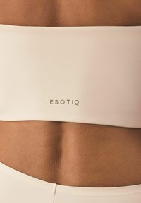 Jasno beżowy top bandeau wykonany z gładkiego materiału o subtelnym połysku, z logo "ESOTIQ" w złotym kolorze z przodu.