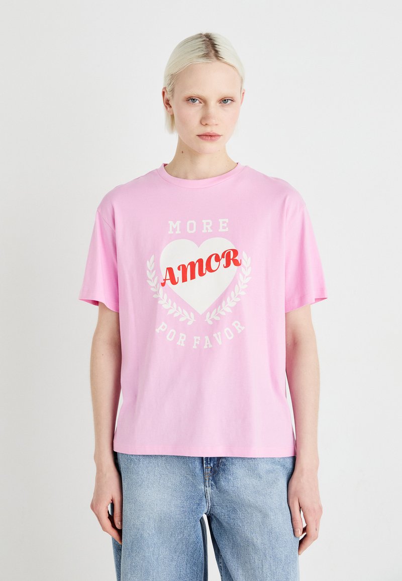 Růžové bavlněné tričko s krátkým rukávem, které má bílé srdce a červený nápis "AMOR" s "MORE" a "POR FAVOR" v světle šedé.