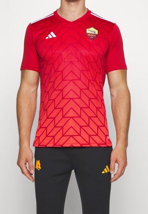 Homme portant un maillot de football rouge de l'AS Roma avec un motif géométrique et un pantalon noir avec des logos jaunes, se tenant contre un fond blanc.