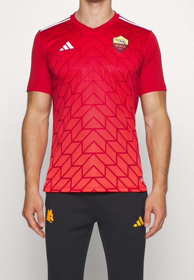 Mies, der ein rotes AS Roma Fußballtrikot mit geometrischem Muster und schwarze Hosen mit gelben Logos trägt, steht vor einem weißen Hintergrund.