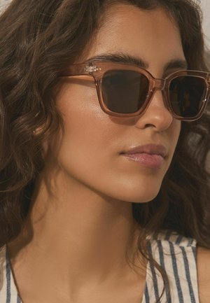 Moteris su garbanotais plaukais, dėvinti permatomus rudus kvadratinius saulės akinius ir dryžuotą be rankovių palaidinę, žiūrinti į dešinę su neutraliu veido išreiškimu.