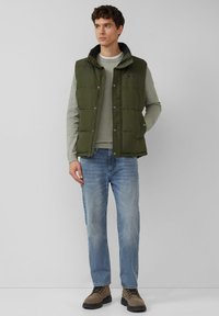 Groene gewatteerde bodywarmer met een hoge kraag, drukknopen en zijzakken, gecombineerd met een lichtgrijs gestreept shirt met lange mouwen en een blauwe spijkerbroek.