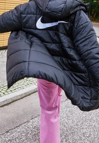 Chaqueta acolchada negra con capucha, con un logo de Nike blanco, llevada sobre pantalones de pana rosa, sobre una acera texturada.
