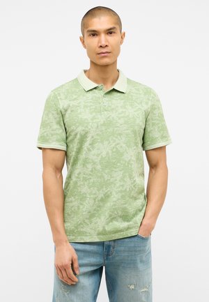 Grünes Poloshirt mit einem strukturierten, blattförmigen Design. Verfügt über einen kontrastierenden, hellgrünen Kragen und eine Knopfleiste. Getragen mit hellen Jeans.