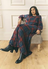 Lång rutig klänning i röd, blå och grön med knapprad; kombinerad med svarta klackstövlar. Modell som sitter på en grå stol.