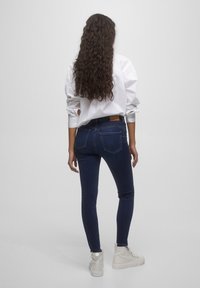 Jeans oscuros y ajustados de cintura alta, con dos bolsillos traseros y una etiqueta de cuero marrón en la cinturilla, combinados con una camisa blanca.