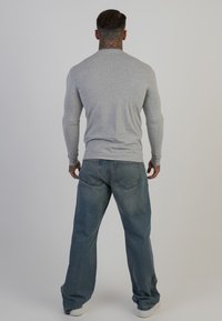 Szürke, hosszú ujjú, testhezálló póló, világoskék, laza farmernadrággal és fehér sportcipőkkel párosítva. A háttér semleges színű.