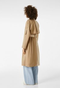 Beżowy trencz z paskiem w talii, naramiennikami i wentylacją z tyłu, zestawiony z jasnoniebieskimi spodniami o szerokich nogawkach. Gładka struktura materiału.