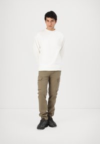 White long-sleeve sweatshirt, khaki cargo pants with multiple pockets, and black textured sneakers, standing against a neutral background.
