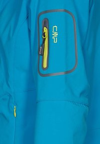 Giacca da esterno blu con tasca con zip verde e dettagli grigi. Texture liscia, design leggero e forma aerodinamica.