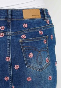 Jeans mini krilo temno modre barve, okrašeno s rožnatim vezenjem in usnjenim oznako "KOROSHI" blizu pasu.