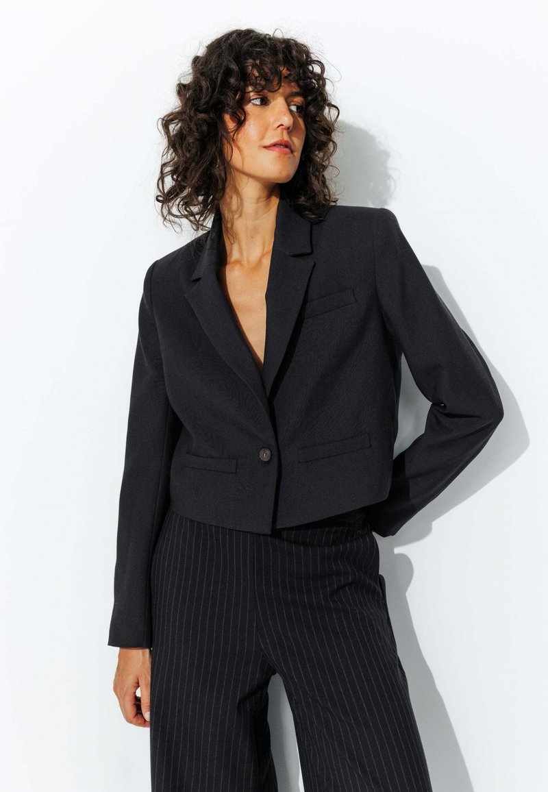 Blazer noir court avec revers crantés, deux poches avant et une fermeture à bouton unique, associé à un pantalon noir à rayures.
