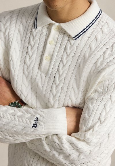 Homme portant un pull en maille torsadée blanche avec un col rayé bleu marine et un "Polo" brodé sur la manche, bras croisés, montrant une montre verte au poignet.