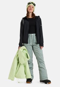 Zwarte ritsjack, zwart shirt, lichtgroene sneeuwbroek en een lichtgroene gewatteerde jas in één hand gehouden. Zwarte laarzen met bruine zolen.