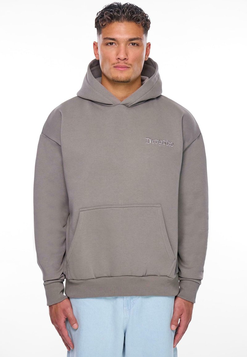 Haut à capuche gris avec une poche avant, présentant un logo brodé discret. Tissu doux et poignets et ourlet côtelés. Porté avec un pantalon en denim clair.