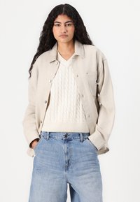 Beige knapplång jacka över en krämfärgad kabelstickad tröja, ihop med ljusblå denimjeans. Texturen är mjuk med en avslappnad passform.