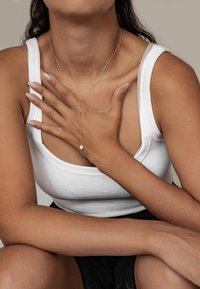 Gouden ketting met een enkele parel pendant, gedragen met een witte geribbelde tanktop. De handen van het model zijn over de borst geplaatst.