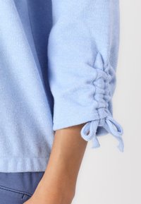 Sweater azul claro com textura canelada, apresentando mangas franzidas atadas com pequenos laços. Material macio e punhos elásticos.