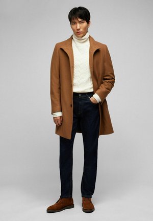 Manteau en laine marron sur un pull à col roulé en maille torsadée blanche, associé à un jean en denim noir et des chaussures en suède marron. Design minimaliste et couleurs classiques.