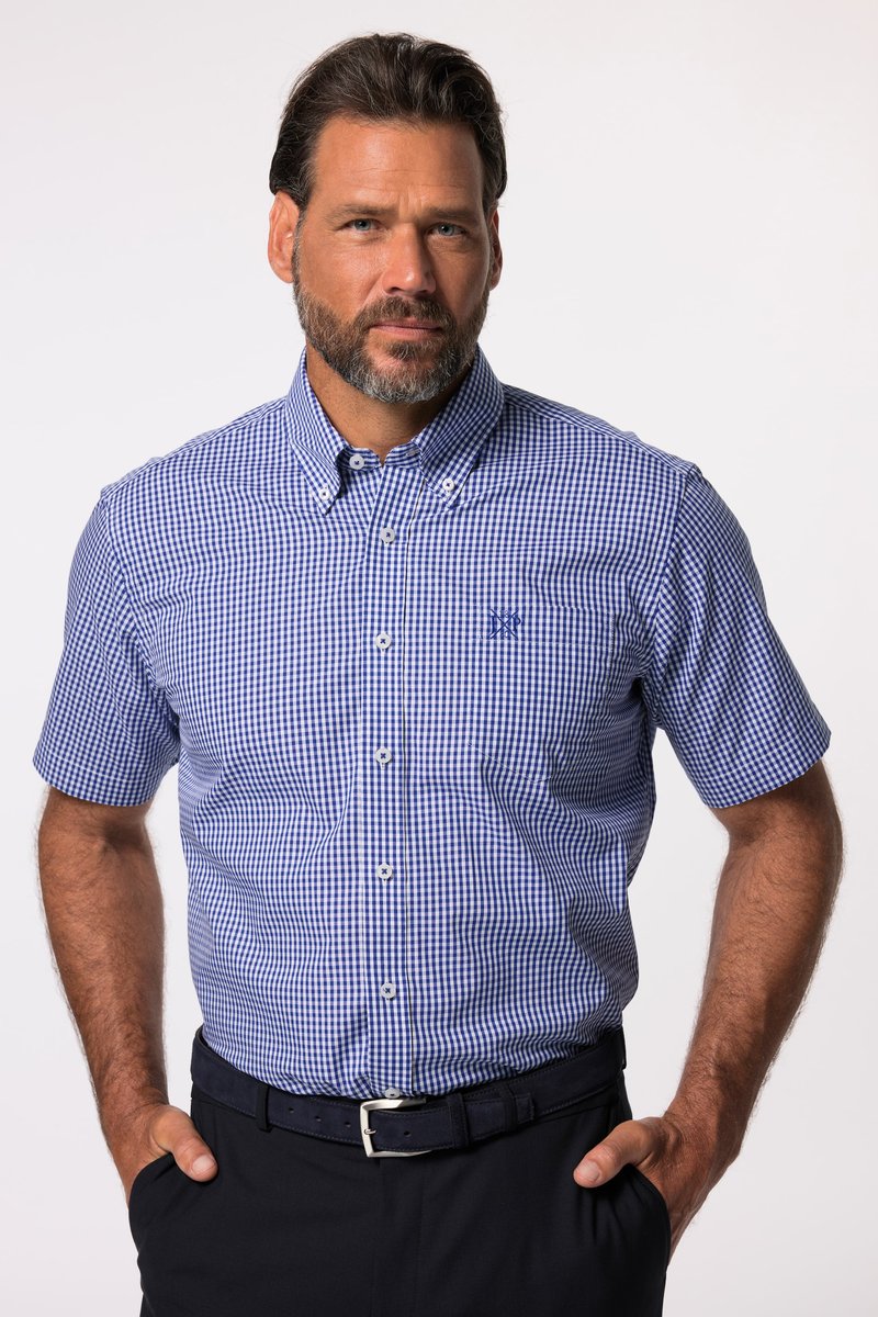 Homme barbu portant une chemise à manches courtes à carreaux bleus et blancs, rentrée dans un pantalon noir avec une ceinture, mains dans les poches.