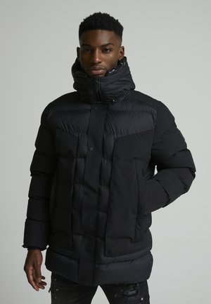 Parka acolchada negra con cuello alto y cierre de cremallera. Presenta botones a presión en el frente y bolsillos laterales. Textura suave, ajuste moderno.