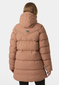 Chaqueta acolchada en marrón claro con capucha, que presenta un diseño acolchado horizontal y un logotipo reflectante en la parte posterior. Textura suave.