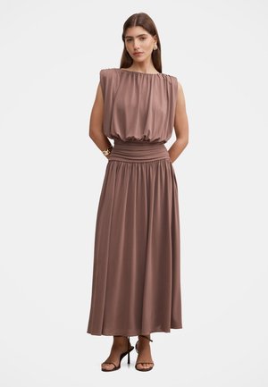 Frau mit langen braunen Haaren, die ein ärmelloses braunes Kleid mit geraffter Taille und schwarze Sandalen mit hohen Absätzen trägt, steht vor einem weißen Hintergrund.