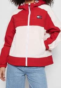 Czerwony i jasnoróżowy wiatrówka z kapturem, zapięciem na zamek, elastycznymi mankietami i przednimi kieszeniami. Posiada naszywkę z logo na klatce piersiowej.