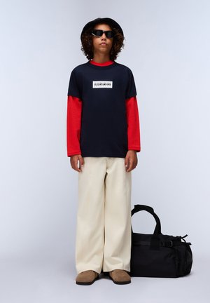 Camiseta navy superpuesta a una camiseta de manga larga roja, pantalones beiges de pierna ancha, zapatos marrones deslizantes y una bolsa de deporte negra. Sombrero negro redondo y gafas de sol.