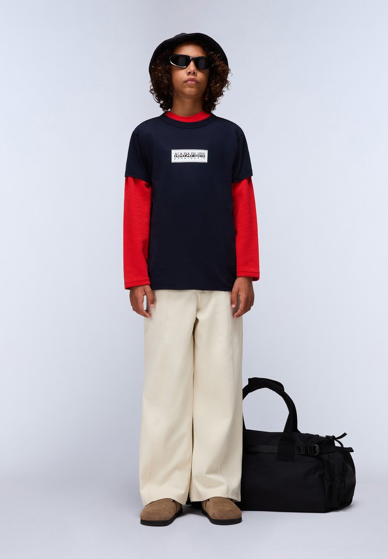 Camiseta navy superpuesta a una camiseta de manga larga roja, pantalones beiges de pierna ancha, zapatos marrones deslizantes y una bolsa de deporte negra. Sombrero negro redondo y gafas de sol.
